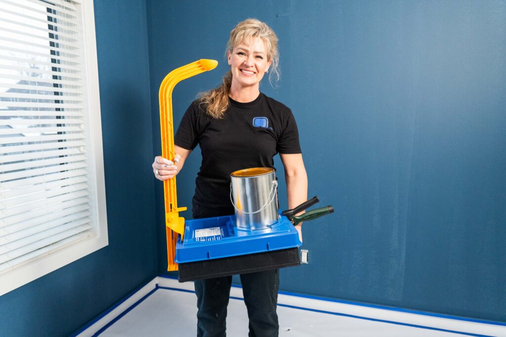 Image of a Karen Wolfe holding a paint tray with a Blue PALOT TRAY PAL lid by a yellow Tray Arm, with all their tools and paint neatly organized together.