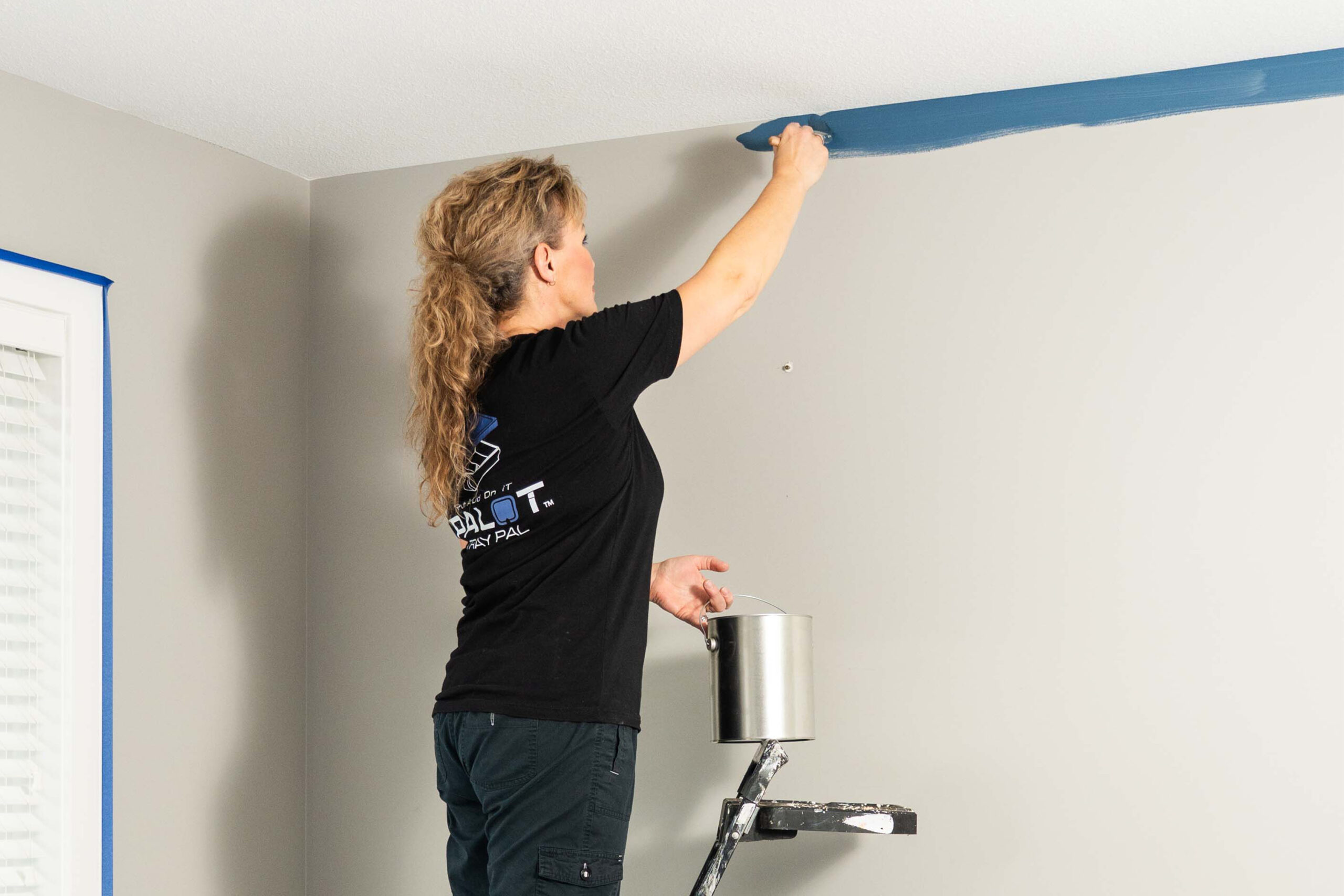 Karen-Wolfe-Painting Karen Wolfe stands on a step ladder holding a can of paint, cutting in the edge of a wall with blue paint.