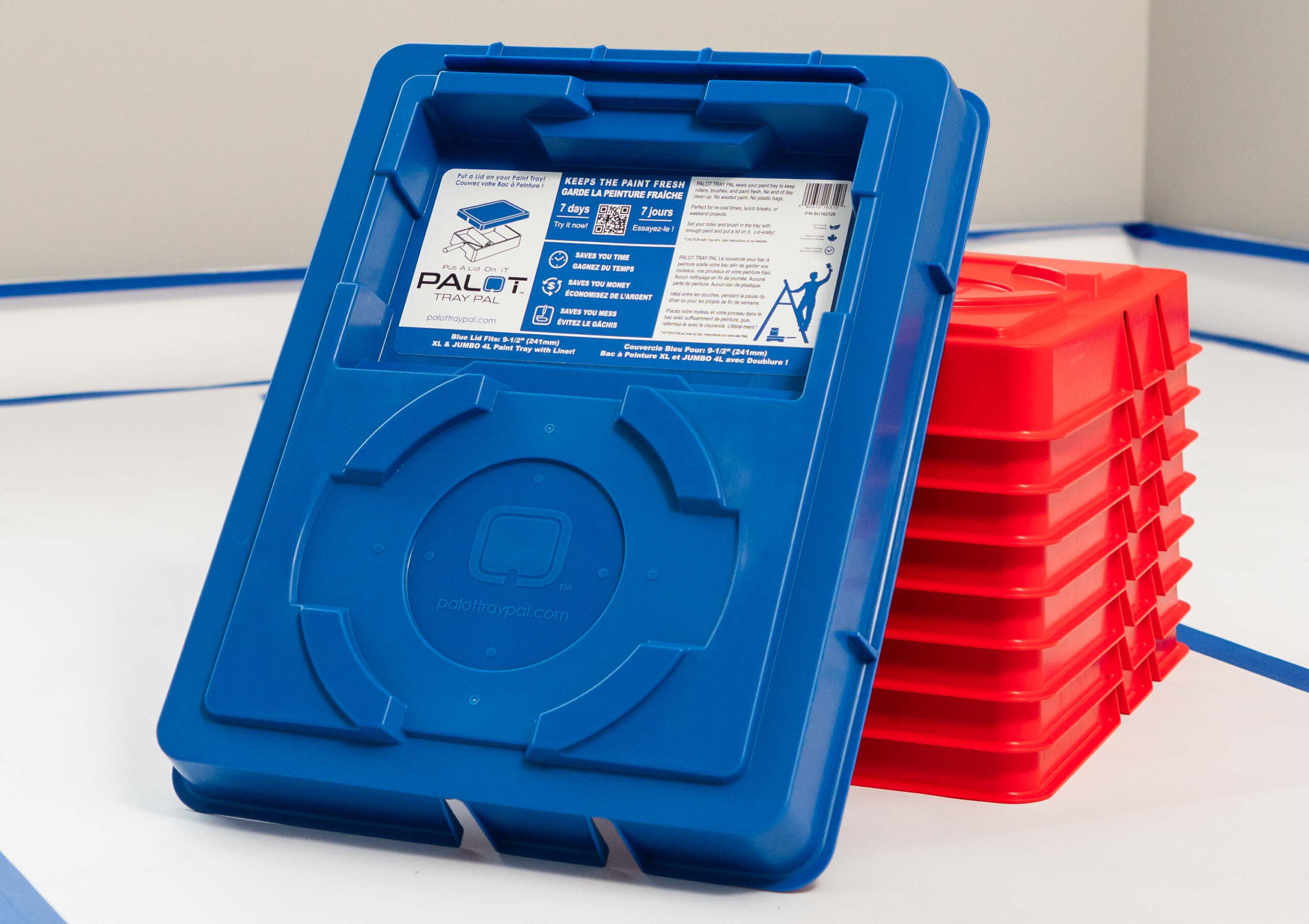 A stack of red PALOT TRAY PAL paint tray lids sits on a floor of a room that is being painted. A Blue PALOT TRAY PAL lid is propped up in front of the stack facing the camera.